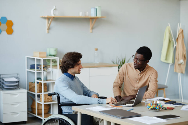 Portrét mladého muže se zdravotním postižením hovořící s kolegou na pracovišti v minimálním kancelářském prostředí, přístupná koncepce pracovních příležitostí - Fotografie, Obrázek