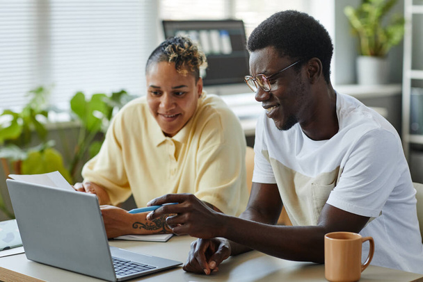 Портрет двох усміхнених чорношкірих людей, які використовують ноутбук разом в ІТ-офісі та носять повсякденний одяг, вказуючи на екран - Фото, зображення