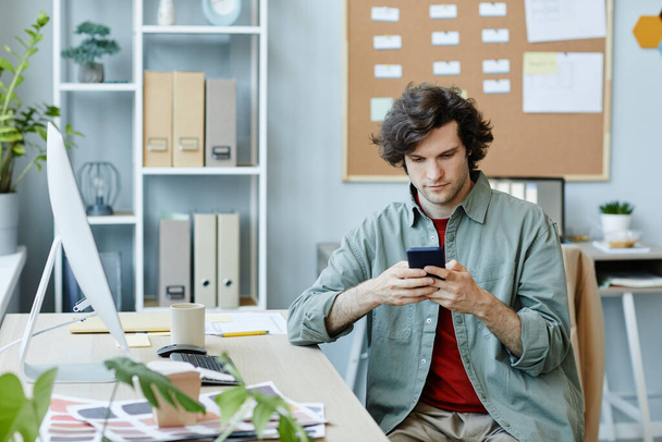 Portret białego młodzieńca korzystającego ze smartfona w miejscu pracy i przewijającego media społecznościowe podczas przerwy - Zdjęcie, obraz