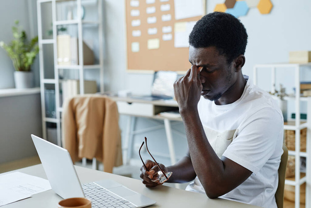 Портрет разочарованного черного мужчины, снимающего очки на рабочем месте и страдающего от головной боли - Фото, изображение
