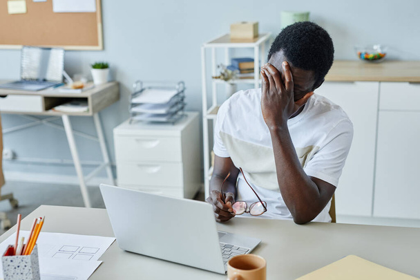 Πορτρέτο του απογοητευμένου μαύρου άνδρα στο χώρο εργασίας που βγάζει γυαλιά και υποφέρει από πονοκέφαλο - Φωτογραφία, εικόνα