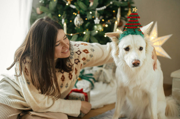 Szczęśliwa kobieta w przytulnym swetrze przytulając ładny biały pies w świątecznym akcesorium na tle stylowej choinki. Zimowe wakacje. Właściciel i zwierzak bawią się w świątecznym pokoju. Wesołych Świąt! - Zdjęcie, obraz