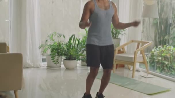 Tilt up shot of athletic man in sportswear jumping rope in living room while doing cardio workout at home - Footage, Video