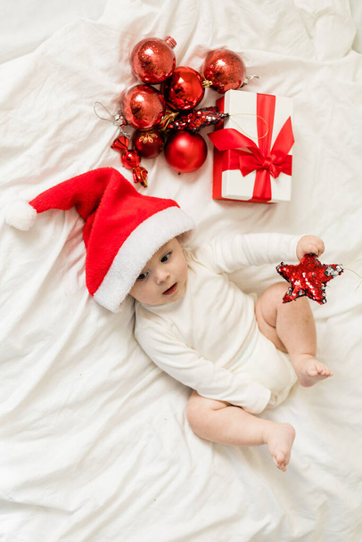 Рождественский малыш в шляпе Санты лежит на кровати с подарочной коробкой и праздничными безделушками вид сверху. - Фото, изображение