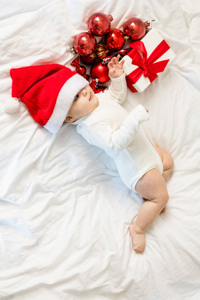 Vánoční batole v Santa klobouk ležící na posteli s dárkovou krabičkou a slavnostní ozdoby horní pohled. - Fotografie, Obrázek