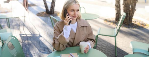 Bild einer stilvollen blonden Frau, die mit dem Handy telefoniert, im Café draußen Kaffee trinkt, das warme Wetter in der Innenstadt genießt, Anrufe auf dem Smartphone entgegennimmt. - Foto, Bild
