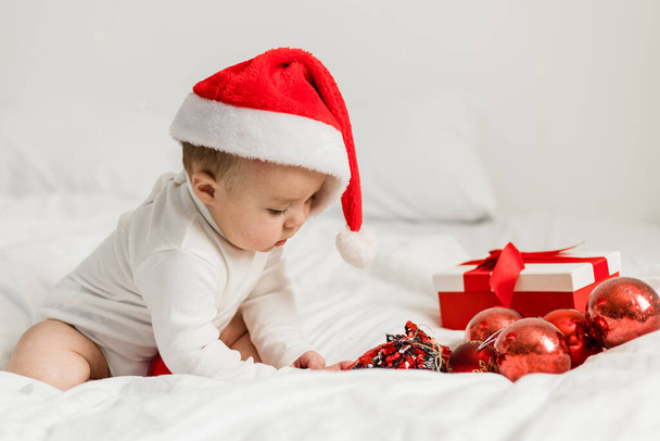 Weihnachtsbaby mit Weihnachtsmütze, Kind hält Christbaumkugel in der Nähe Geschenk-Box über Weihnachtsbeleuchtung - Foto, Bild
