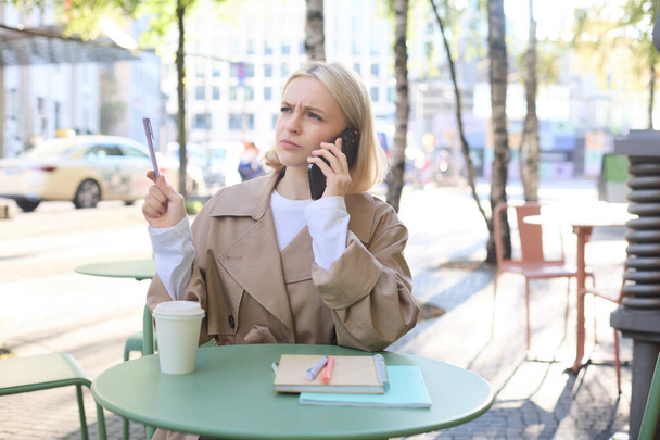 Εικόνα της γυναίκας με συγκεχυμένο, σοβαρό πρόσωπο, απαντά στο τηλέφωνο, εργάζεται σε cafe σε εξωτερικούς χώρους, πίνοντας καφέ, συνοφρύωμα με στυλό στο χέρι. - Φωτογραφία, εικόνα