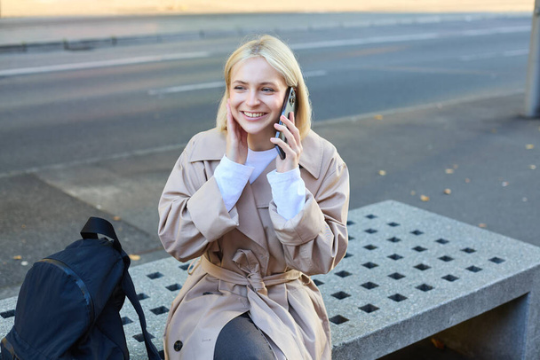 Image of young blond woman sitting on street bench with backpack, talking on mobile phone, answers a call and smiling while chatting with someone. - Photo, Image