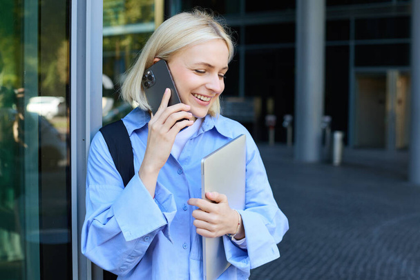 Mladá moderní žena, zaměstnankyně, stojí v blízkosti kancelářské budovy, zvedá telefon, mluví, drží notebook a batoh, usmívá se, zatímco mají pěkný, přátelský chat. - Fotografie, Obrázek