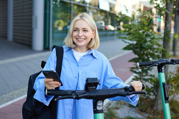 Ritratto di giovane donna sorridente, sblocca scooter elettrico, scansiona il codice qr con l'app per smartphone per sbloccare il suo giro, in piedi sulla strada vicino all'edificio. - Foto, immagini