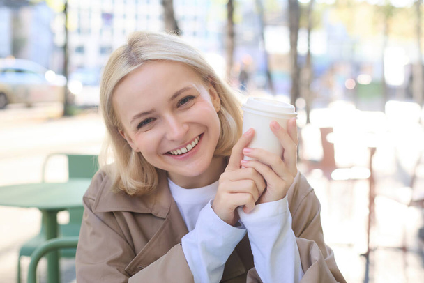 Primer plano retrato de una mujer rubia sonriente, estudiante universitaria, sosteniendo una taza de café, calentando sus manos en el frío día de otoño, sentada en la cafetería al aire libre, riéndose de la cámara. - Foto, imagen