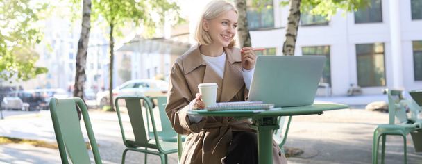 Зображення молодої блондинки сучасної жінки, яка сидить з ноутбуком зовні в кафе, п'є кавовий напій і працює над проектом, використовуючи комп'ютер. - Фото, зображення