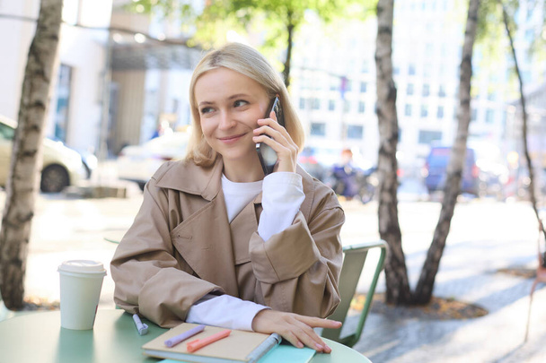 Gülümseyen sarışın kadının yakın portresi, kafenin dışındaki masada oturuyor, cep telefonuyla konuşuyor, mutlu, canlı sohbet ediyor.. - Fotoğraf, Görsel