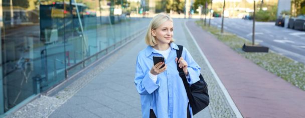 Portrait of blond young woman, using navigation map app on smartphone, walking along street, using directions on phone, holding backpack. - Photo, Image