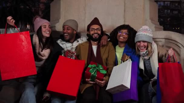 Ryhmä kansainvälisiä ystäviä ostoskasseineen ja joululahjoineen hymyilemässä, nauramassa ja katselemassa kameraa. Onnellisia kavereita juhlimassa joulua. Nuoret aikuiset nauttivat joulu-illasta - Materiaali, video