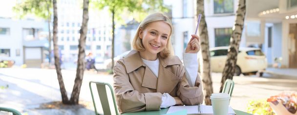 Portret van een jonge studente, die aan haar documenten werkt, buiten huiswerk maakt in café, koffie drinkt en essay schrijft, marker vasthoudt, glimlacht naar de camera. - Foto, afbeelding