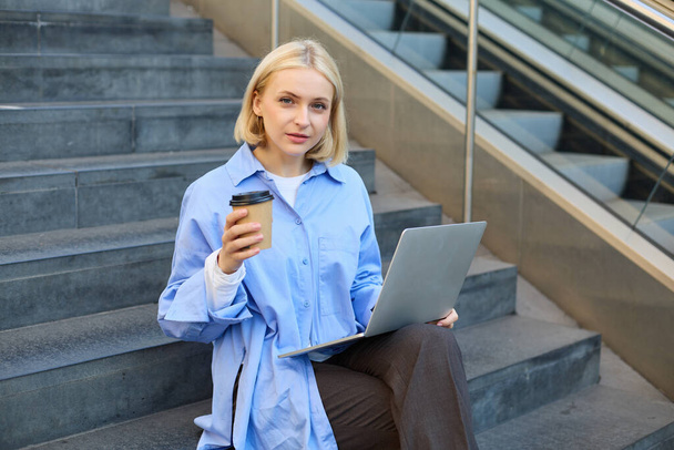 Ritratto di giovane donna urbana seduta sulle scale con tazza di caffè da asporto e che lavora su laptop, studia all'aperto all'aria aperta, freelance. - Foto, immagini