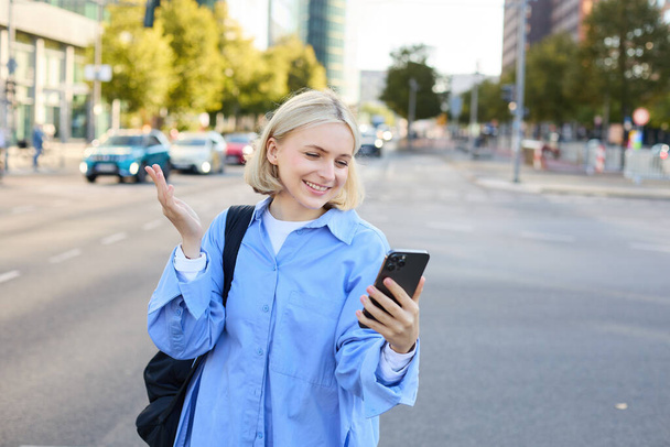 Portret młodej pracowniczki biurowej, kobieta spacerująca ulicą, rozmawiająca przez telefon, patrząca na smartfona i pokazująca smth za nią, czaty wideo, łączy się z rozmową online za pośrednictwem aplikacji mobilnej. - Zdjęcie, obraz