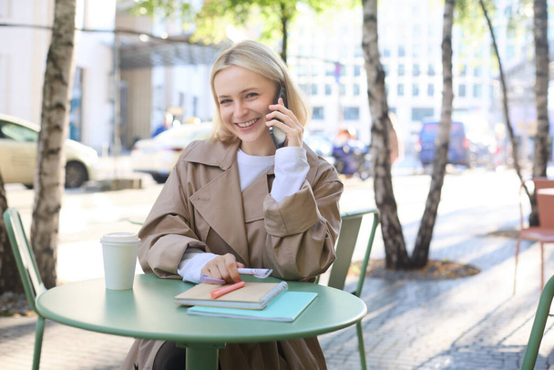 Elämäntapa ja kaupunkilaisten käsite. Nuori hymyilevä tyttö, opiskelija istuu kahvilassa ulkona, puhuu matkapuhelimella, juttelee ystävän tai työtoverin kanssa puhelimessa, opiskelee tai tekee läksyjä. - Valokuva, kuva
