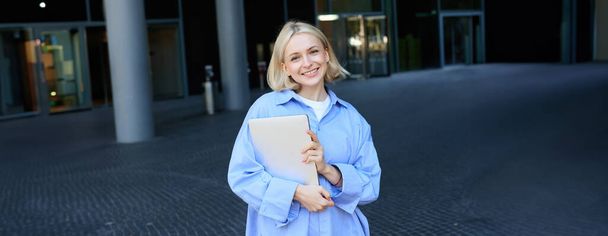 Porträt einer jungen blonden Frau, Studentin, die mit Notizbüchern und Dokumenten in der Nähe ihres Campus steht, blaues Hemd trägt und in die Kamera lächelt. Bildungskonzept - Foto, Bild