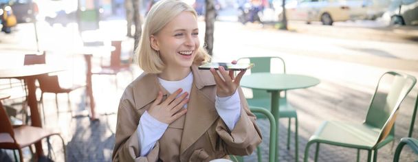 Портрет счастливой молодой женщины, сидящей в кафе и записывающей голосовое сообщение, держащей мобильный телефон у губ и говорящей в микрофон, проводящей время в кафе, пьющей кофе и чате. - Фото, изображение