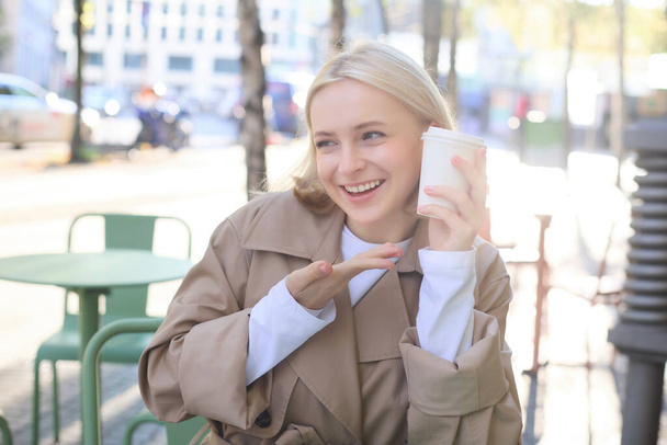 Portrait of smiling blond woman, pointing finger at her cup of coffee, orders takeaway, sits in street cafe, shows her favourite drink. - Photo, Image