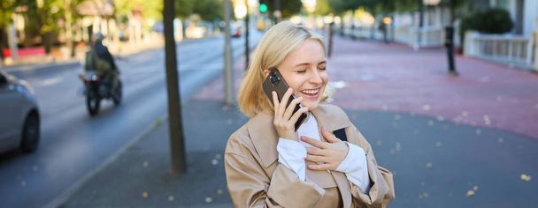 Κοντινό πλάνο πορτρέτο της όμορφης νεαρής γυναίκας, ξανθό κορίτσι που περπατά στο δρόμο με το κινητό τηλέφωνο, κουβεντιάζοντας με το φίλο, έχει χαρούμενη έκφραση προσώπου, ενώ μιλάμε πάνω από το κινητό τηλέφωνο στο δρόμο της. - Φωτογραφία, εικόνα