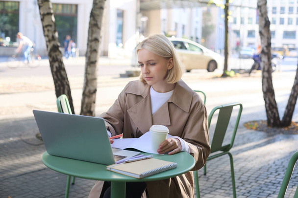 Изображение молодой современной женщины, сидящей в кафе на открытом воздухе, пьющей кофе или чай, смотрящей на ноутбук, работающей над проектом, фрилансером, делающей заметки в блокноте. Концепция образа жизни - Фото, изображение