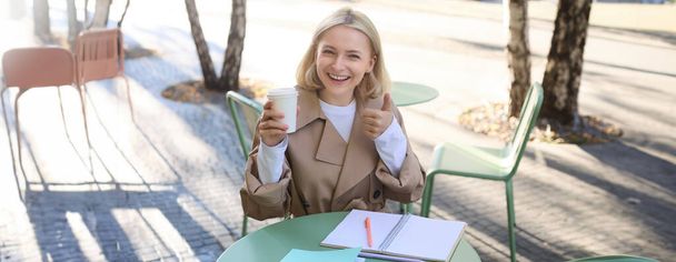 Lächelnde, fröhliche Frau, die im Straßencafé ihren Kaffee trinkt, in Notizbücher schreibt, Hausaufgaben macht, Notizen macht. Studenten- und Lifestylekonzept. - Foto, Bild