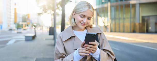 Beeld van een jonge moderne vrouw, studente die op straat loopt en naar haar mobiele telefoon kijkt, glimlacht en er gelukkig uitziet. Kopieerruimte - Foto, afbeelding