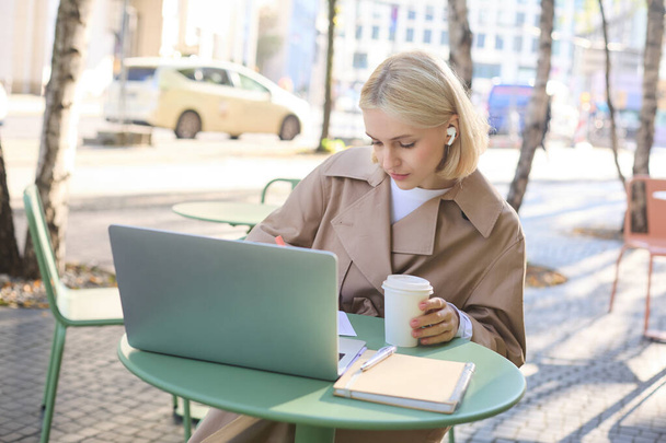 Ritratto di giovane donna bionda con computer portatile, seduta per strada, che beve caffè in un caffè all'aperto, lavora, indossa cuffie senza fili, fa i compiti, freelance. - Foto, immagini