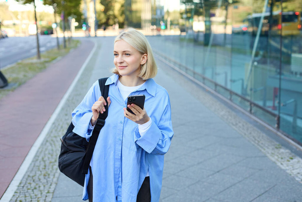 Portrait of blond young woman, using navigation map app on smartphone, walking along street, using directions on phone, holding backpack. - Photo, Image