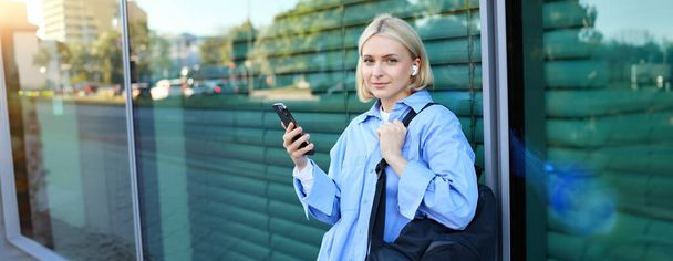 Retrato de estilo de vida de la joven modelo sonriente, estudiante con mochila, esperando a alguien en la calle, de pie al aire libre con teléfono inteligente. - Foto, Imagen