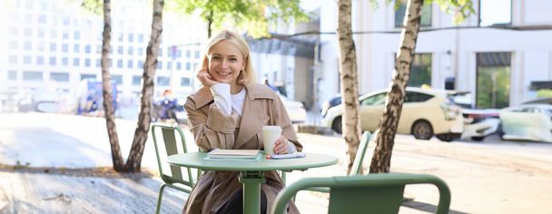 Koncept městského životního stylu. Mladá krásná žena sedí ve venkovní kavárně, přemýšlí o životě ve městě, pije kávu a usmívá se na ulici. - Fotografie, Obrázek