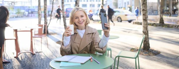 Estilo de vida de chica rubia con estilo con teléfono inteligente, beber café, posando con su chai para las redes sociales, la creación de contenido en el teléfono, tomar selfie. - Foto, Imagen