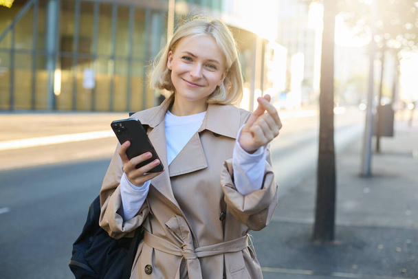 Piękna młoda modelka w płaszczu, pokazująca gest serca palcami, trzymająca smartfona i uśmiechnięta do aparatu, stojąca na pustej, słonecznej ulicy. - Zdjęcie, obraz