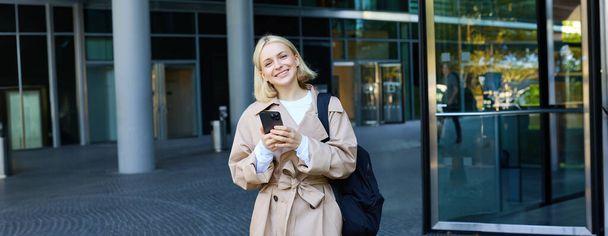 Портрет молодой стильной женщины, гуляющей по центру города со смартфоном, использующей мобильное приложение на открытом воздухе, ожидающей кого-то, идущей на работу. - Фото, изображение
