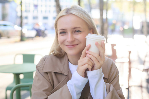 Primo piano di donna bionda allegra, con in mano una tazza di caffè, bere tè in strada caffè all'aperto, sorridente con espressione felice del viso. - Foto, immagini