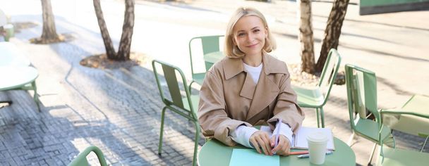 Portrait de jeune femme européenne souriante, travaillant à l'extérieur, écrivant des notes dans un carnet, buvant du café à emporter et regardant avec bonheur la caméra. Concept de style de vie - Photo, image