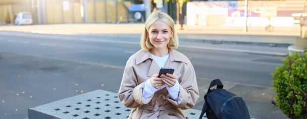 Venkovní záběr mladé blondýny na ulici, sedící na lavičce s mobilním telefonem, usmívající se do kamery, čekající na kamarádku mimo budovu, která si dává pauzu. - Fotografie, Obrázek