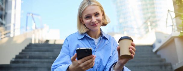 Ritratto di bella donna bionda, studentessa sorridente che beve caffè, seduta sulle scale con smartphone, guardando felice la macchina fotografica. - Foto, immagini