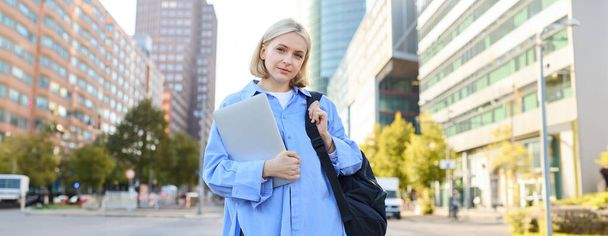Porträt einer jungen selbstbewussten Frau, College-Studentin mit Rucksack und Laptop, auf dem Weg zum Unterricht, steht draußen auf einer leeren Straße mit großen Gebäuden dahinter. - Foto, Bild