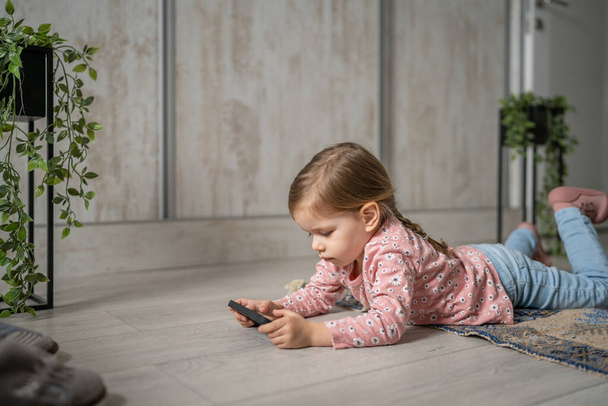 une fille caucasien enfant tout-petit tenir smartphone téléphone mobile à la maison jouer à des jeux vidéo enfance et grandir technologie addiction concept utiliser application pour la navigation en ligne ou regarder la vidéo - Photo, image
