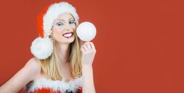 Beautiful blonde girl in Santa hat with white Christmas ball in hand. Smiling woman with festive makeup with New year tree toy. Christmas holiday. Christmas sale. Discount. Copy space for advertising - Photo, Image