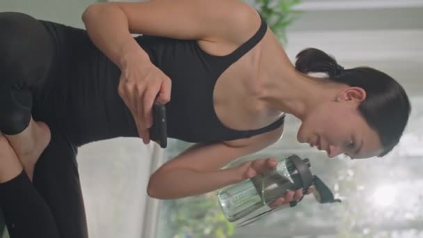 Вертикальный снимок молодой женщины в спортивной одежде, просматривающей социальные сети на смартфоне и питьевой воде во время отдыха после тренировки дома - Кадры, видео