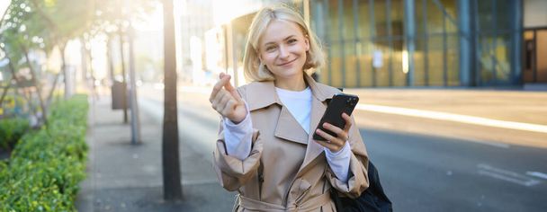 Charmant modèle jeune femme en trench coat, montrant un geste de cœur avec les doigts, tenant smartphone et souriant à la caméra, debout sur la rue ensoleillée vide. - Photo, image