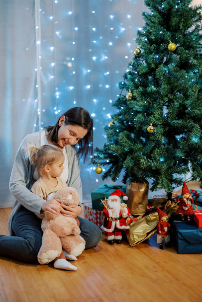 La petite fille avec un ours en peluche s'assoit sur le genou de sa mère sur le sol et regarde les cadeaux sous l'arbre. Photo de haute qualité - Photo, image