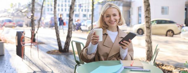 Портрет белокурой улыбающейся женщины, сидящей в кафе, наслаждающейся чашкой кофе на вынос, использующей мобильный телефон, работающей на смартфоне, блоггер социальных сетей, делающий пост в Интернете. - Фото, изображение