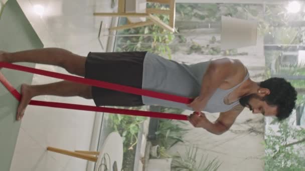 tiro vertical de homem muscular em sportswear fazendo exercício em linha reta com banda de resistência durante o treinamento em casa - Filmagem, Vídeo